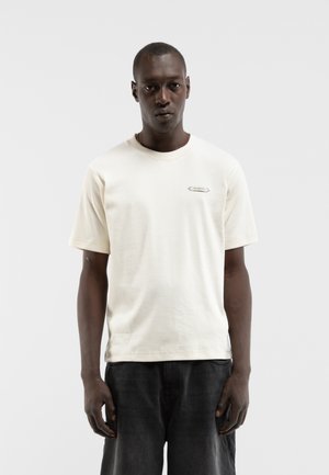 Homme debout, portant un t-shirt uni beige clair à manches courtes et un pantalon foncé sur un fond gris clair uni.