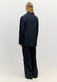 Marineblaues Nadelstreifen-Anzugset mit einem langen Blazer mit einem einzelnen Rückenschlitz und weiten Hosen mit Cargo-Taschen. Glatter, maßgeschneiderter Stoff.
