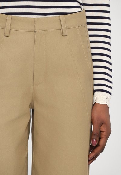 Pantaloni beige su misura con una texture liscia, caratterizzati da dettagli cuciti e tasche laterali. Una maglietta a righe a maniche lunghe è parzialmente visibile.
