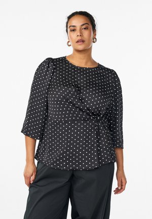 Žena v čiernej blúzke s bodkami s rukávmi do polovice ramena a čiernych nohaviciach stojaca pred jednoduchým bielym pozadím.