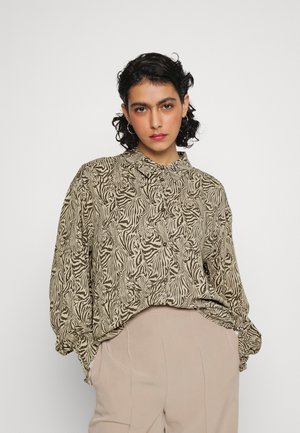 Camisa bege de botão com um padrão negro ondulado, com colarinho e corte solto, combinada com calças claras. Textura suave.