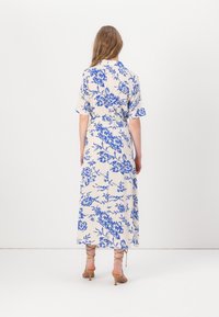 Žena stojící zády, oblečená v bílé midi šatech s modrým květinovým vzorem a hnědými sandály na podpatku, na bílém pozadí.