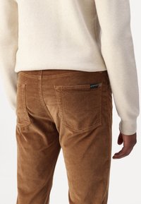 Pantalons en velours côtelé marron avec une texture à côtes verticales ; comportent deux poches arrière et une petite étiquette de marque près de la taille. Un pull est visible au-dessus.