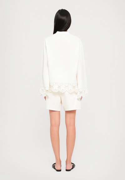 Woman with long dark hair wearing a white long-sleeve top with lace trim and white shorts, standing facing away on a plain background.