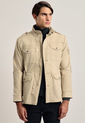 Hombre vestido con una chaqueta beige abotonada y con cremallera sobre un jersey de cuello alto negro y pantalones negros, mirando hacia la derecha contra un fondo liso.