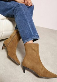 Brūni zamšādas potīšu zābaki ar vērsta pirksta formu un 5 cm augstu stiletto papēdi. Iekļauts sānu rāvējslēdzis un gluda virsma. Valkāti kopā ar zilo džinsu.