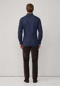 Man staat facing een eenvoudige muur in een donkerblauw gestreept overhemd met lange mouwen, donkerbruine broek en bruine schoenen.