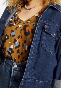 Chemise en denim oversize superposée à une chemise marron ornée d'un motif léopard noir, orange et bleu. Accent en métal argenté sur le jean.