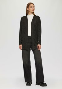 Schwarze Langarmstrickjacke mit strukturiertem Muster, offene Front, getragen über einem weißen Rollkragenpullover und schwarzen, weit geschnittenen Jeans. Voluminöse schwarze Stiefel.