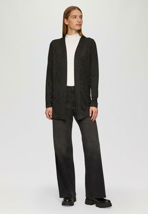 Black, long-sleeve cardigan with a textured pattern, open front, worn over a white turtleneck and black wide-leg jeans. Chunky black boots.