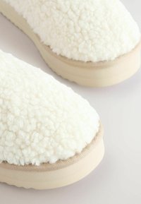 Chaussons blancs en peluche avec une tige texturée et douce, des accents en tissu beige, et une semelle durable de couleur claire. Forme lisse et arrondie.