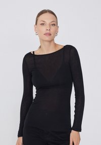 Haut noir à manches longues en tissu transparent, doté d'un col bateau et d'une silhouette ajustée. Texture subtile avec transparence sur la partie supérieure du corps.