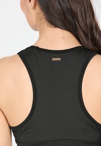Zwarte sportieve tanktop met een gestructureerde stof, racerback ontwerp en een klein gouden logo-detail bij de hals. Gladde afwerking.