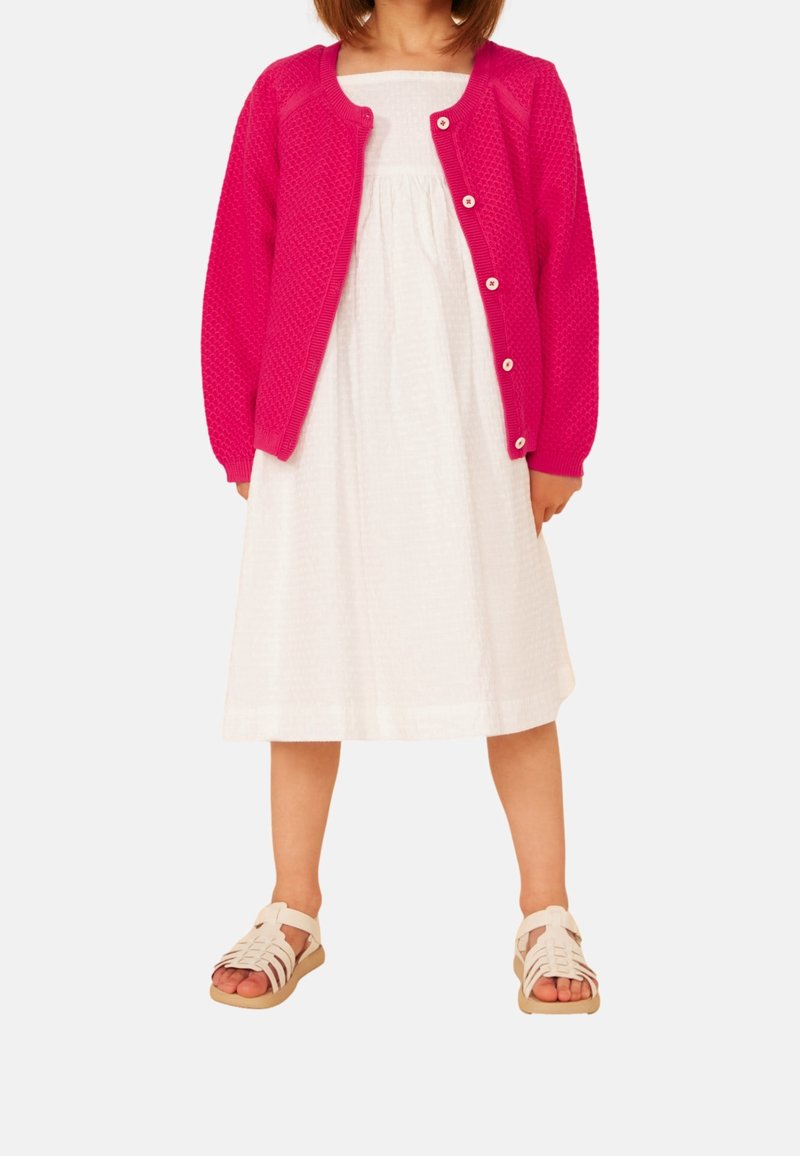 Enfant portant une robe blanche sans manches, un cardigan boutonné rose vif et des sandales blanches, debout les pieds légèrement écartés.