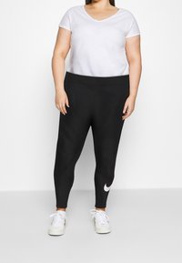 Kvinna iklädd svarta Nike leggings med vit swoosh på vänster vad, vit kortärmad tröja och vita sneakers, som står mot en enfärgad bakgrund.