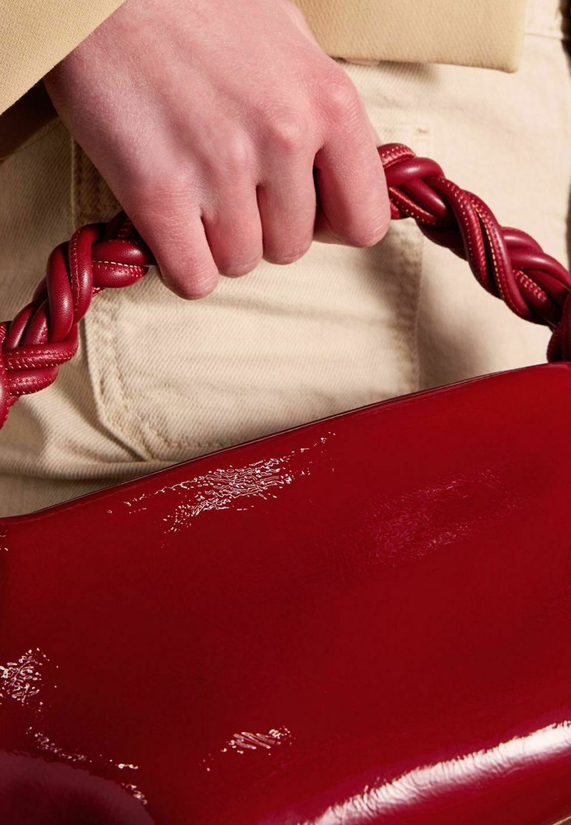 Red handbag with a glossy finish and a braided top handle, held by a hand. Background features light-colored pants.