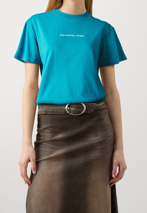 Mujer que lleva una camiseta turquesa con el texto "COLOURFUL REBEL" metida en una falda marrón descolorida de talle alto con cinturón de hebilla ovalada.