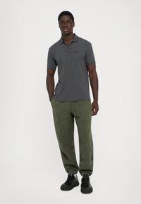 Man stands wearing a dark grey polo shirt and olive green jogger pants with black sneakers against a plain light background.