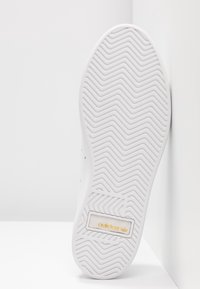 Vit sneakersula med texturerad zigzag-mönster, med en Adidas-logotyp i guld på ett beige accentområde. Slät, ren design överlag.