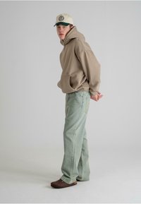 Beige hette med frontlomme, lysegrønne rettskårne jeans, brune sko og en to-farget caps med logo. Modell står sidelengs.