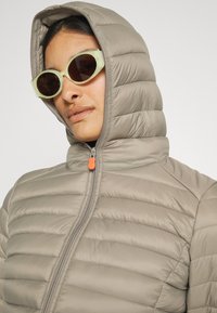 Lichtgrijze pufferjack met een hoge kraag en capuchon, voorzien van horizontale stiksels, een rits en een oranje ritstrekker.