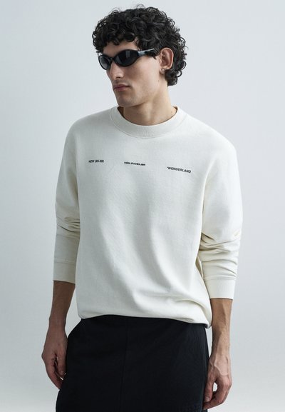 Hvid sweatshirt med trykt tekst i sort, med rund halsudskæring og lange ærmer. Kombineret med sorte solbriller og mørke bukser.