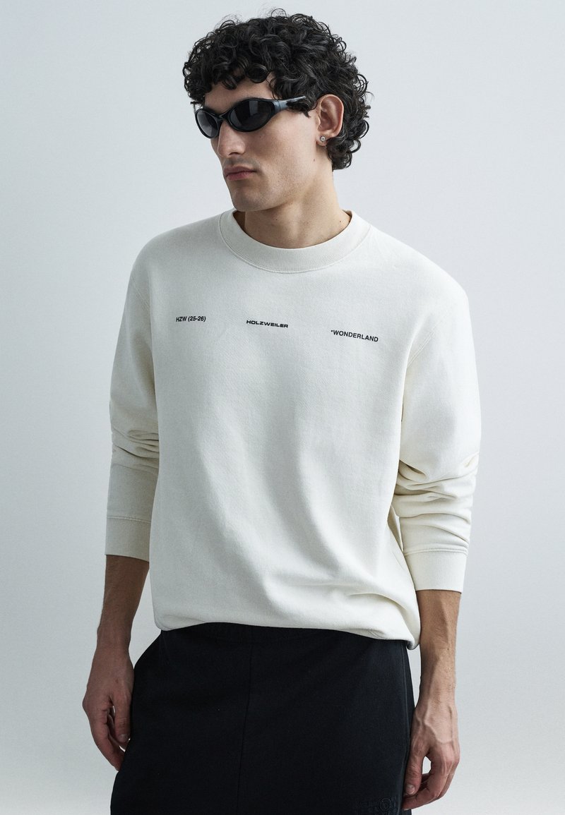 Biela mikina s čiernym potlačeným textom, vybavená okrúhlym výstrihom a dlhými rukávmi. Kombinovaná s čiernymi slnečnými okuliarmi a tmavými nohavicami.