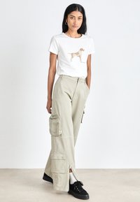 Weißes Baumwollt-Shirt mit einem Grafikdruck eines braunen Hundes, kombiniert mit hellgrünen Cargo-Hosen mit Reißverschlusstaschen und mehreren Taschen. Schwarze Sneakers.