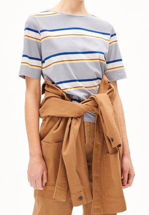 Henkilö, jolla on harmaa lyhythihainen paita, jossa on sinisiä ja keltaisia raitoja, ja ruskea takki solmittuna vyötärön ympäri.