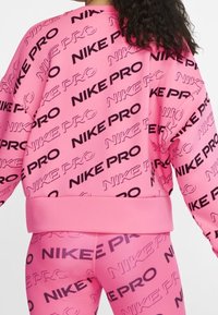 Felpa rosa e leggings con testo ripetuto "NIKE PRO" in nero. Il tessuto ha una texture liscia e un design aderente.