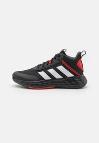 Chaussure de sport noire avec des rayures blanches et des accents rouges. Présente un matériau en maille, une semelle épaisse et texturée, ainsi qu'un détail de talon contrastant.
