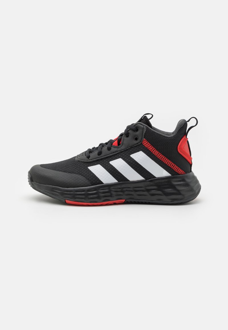 Chaussure de sport noire avec des rayures blanches et des accents rouges. Présente un matériau en maille, une semelle épaisse et texturée, ainsi qu'un détail de talon contrastant.