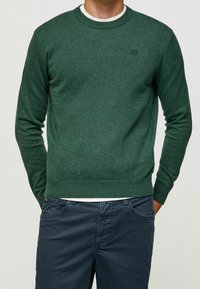 Groen gebreid sweater met ronde hals en geribbelde manchetten. Draagt over een wit shirt, gecombineerd met donkerblauwe broek. Subtiele logo-detail op de mouw.