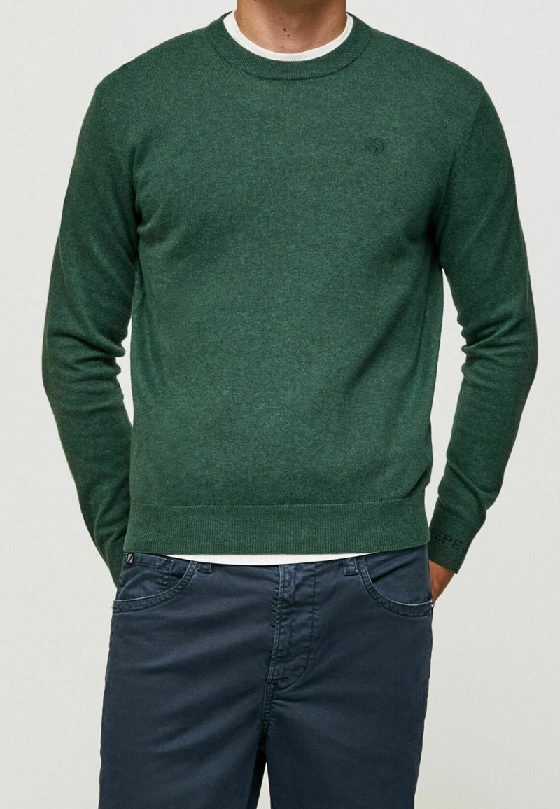Groen gebreid sweater met ronde hals en geribbelde manchetten. Draagt over een wit shirt, gecombineerd met donkerblauwe broek. Subtiele logo-detail op de mouw.