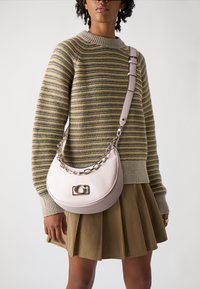 Borsa in pelle beige dalla forma arrotondata, con una tracolla in catena argentata e un logo prominente. Indossata sopra un maglione a righe e una gonna plissettata.