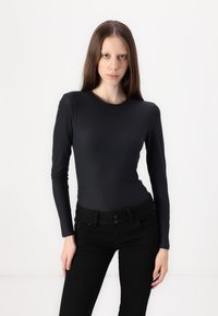 Junge Frau mit langen, glatten Haaren, die ein eng anliegendes schwarzes Langarmoberteil und schwarze Hosen trägt, im Stehen vor einem schlichten weißen Hintergrund.