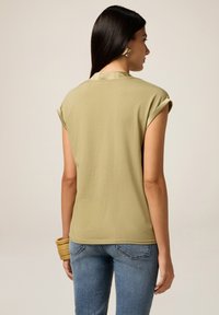 Donna che indossa una t-shirt beige con le maniche arrotolate, jeans blu, orecchini quadrati dorati e braccialetti impilati dorati, di spalle contro uno sfondo neutro.