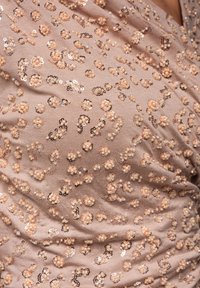 Beżowa tkanina z kwiatowym wzorem z cekinów w różnych odcieniach brzoskwini i złota, tworząca teksturę i wizualne zainteresowanie. Gładka, lekko układająca się fałdami.