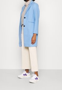 Lichtblauwe blazer met twee knopen, gecombineerd met een grijze coltrui en crèmekleurige bredere broek. Witte sneakers met paarse accenten.