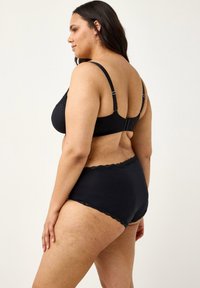 Plus-size kvinde iført sort bh og matchende højtaljet undertøj med blonderkant, stående i profil mod en ensfarvet baggrund.