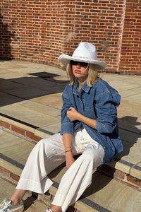 Chaqueta de mezclilla con cierre de botones, pantalones anchos blancos y un sombrero de sol blanco de fibra trenzada, sentado en escalones de piedra con una pared de ladrillo texturizada detrás.