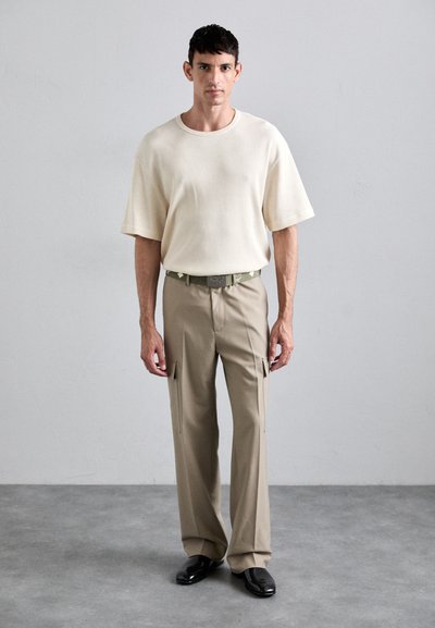 Beige kortermet strikket skjorte kombinert med lyse khaki cargo-bukser med frontlommer; modell iført svarte sko. Minimalistisk design.