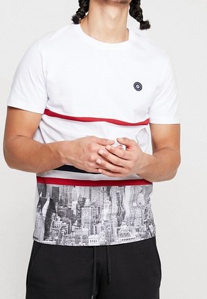 Mand iført en hvid T-shirt med røde striber og et sort-hvidt bylandskabsprint på den nederste halvdel, kombineret med sorte bukser.