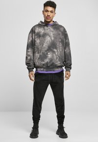Siva kapuljača s tie-dye uzorkom i kenguruhu čepom te crne joggerice. Komplet ima opušteni kroj i meku teksturu. Crne tenisice zaokružuju izgled.