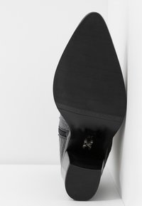 Vue de la semelle d'une botte noire à bout pointu avec une tige texturée et un talon bloc, taille 37, positionnée contre un angle blanc.