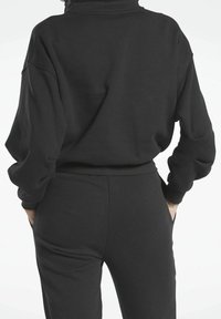 Osoba oblečená v černém dlouhém rukávu a odpovídajících černých kalhotách, stojí s rukama v kapsách, otočená zády k fotoaparátu.