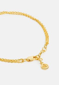 Armbands av guldmetall med dubbla kedjelänkar, utrustad med ett lås och en rund berlock som visar en stiliserad bokstav.