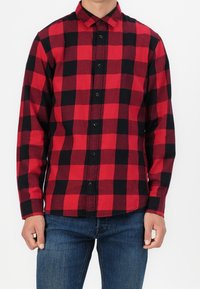 Hombre vistiendo una camisa de cuadros rojos y negros de botones de manga larga, combinada con jeans azules, frente a un fondo blanco liso.
