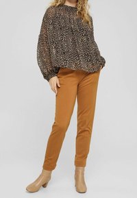 Blusa nera con stampa leopardata beige, maniche ampie e scollatura arricciata, abbinata a pantaloni marroni attillati e stivali con tacco beige alla caviglia.