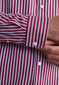 Rot-weiß gestreiftes Hemd mit langen Ärmeln. Verfügt über einen weißen Knopf und eine kontrastierende Streifentextur entlang der Bündchen.